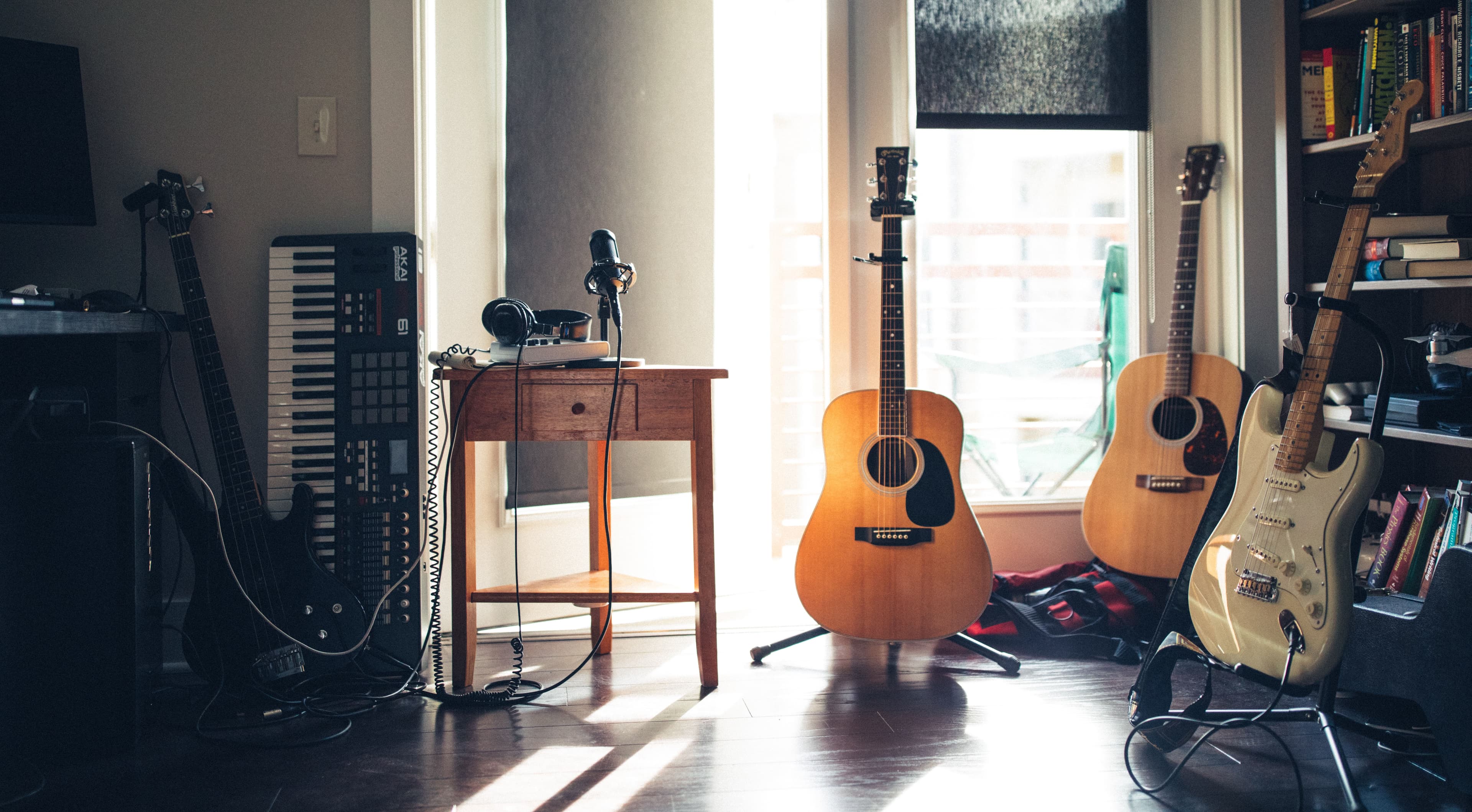 A room with a bass guitar, a keyboard, recording equipment, 2 acoustic guitars, and an electric guitar beside a bookcase full of books